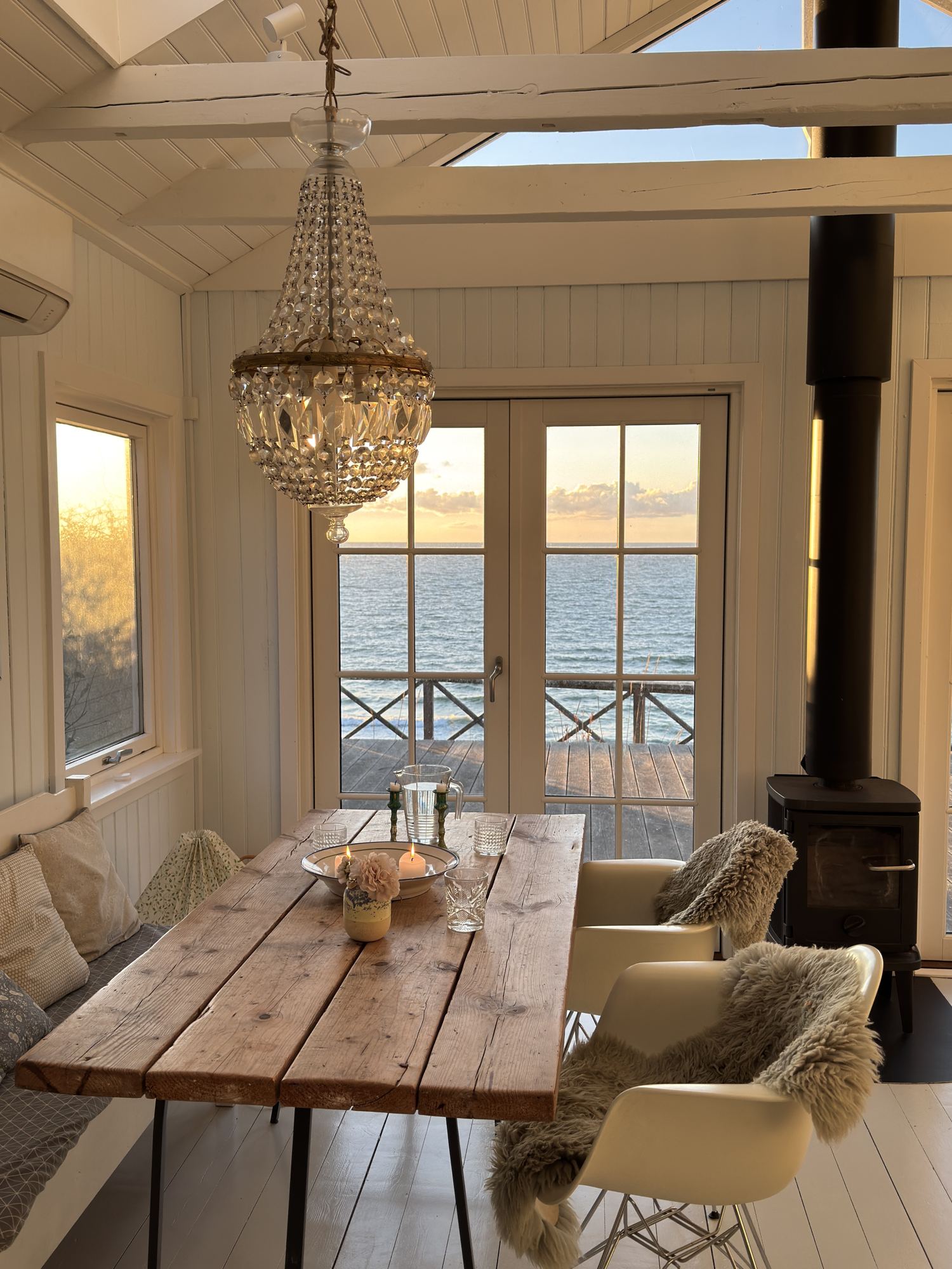 Wooden dining table under a crystal chandelier in a white summer house, with the sea at sunset through French doors