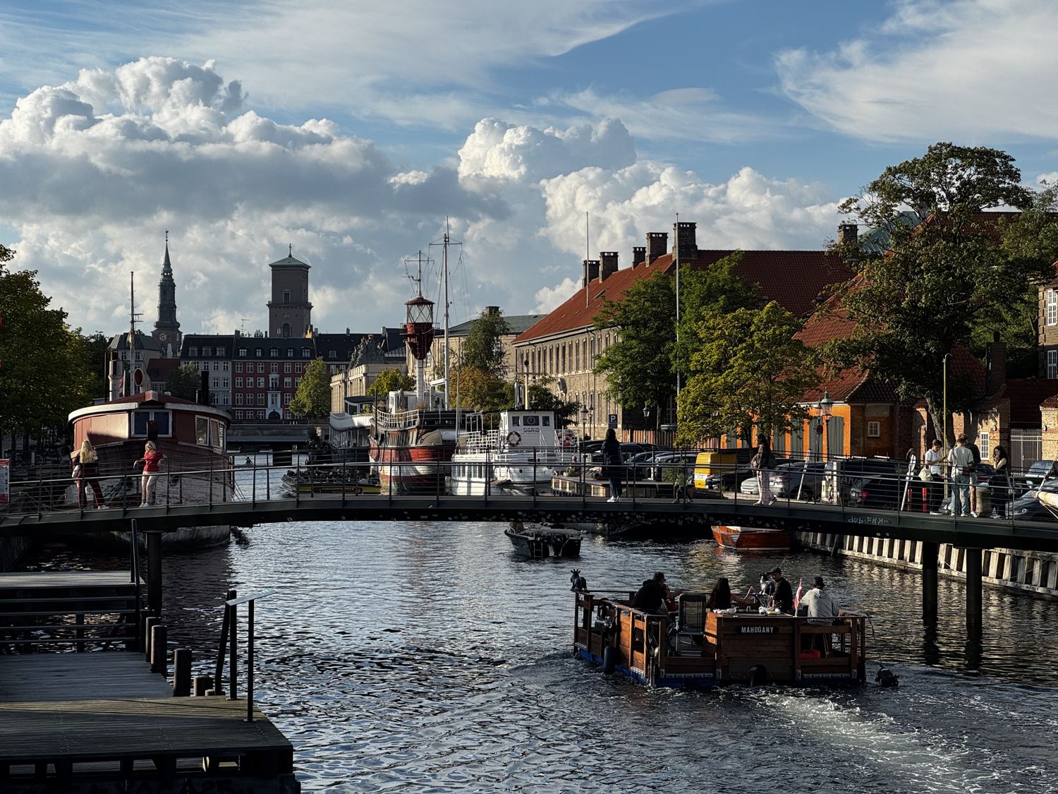 A wooden boat on a Copenhagen canal under a footbridge, with church spires behind
